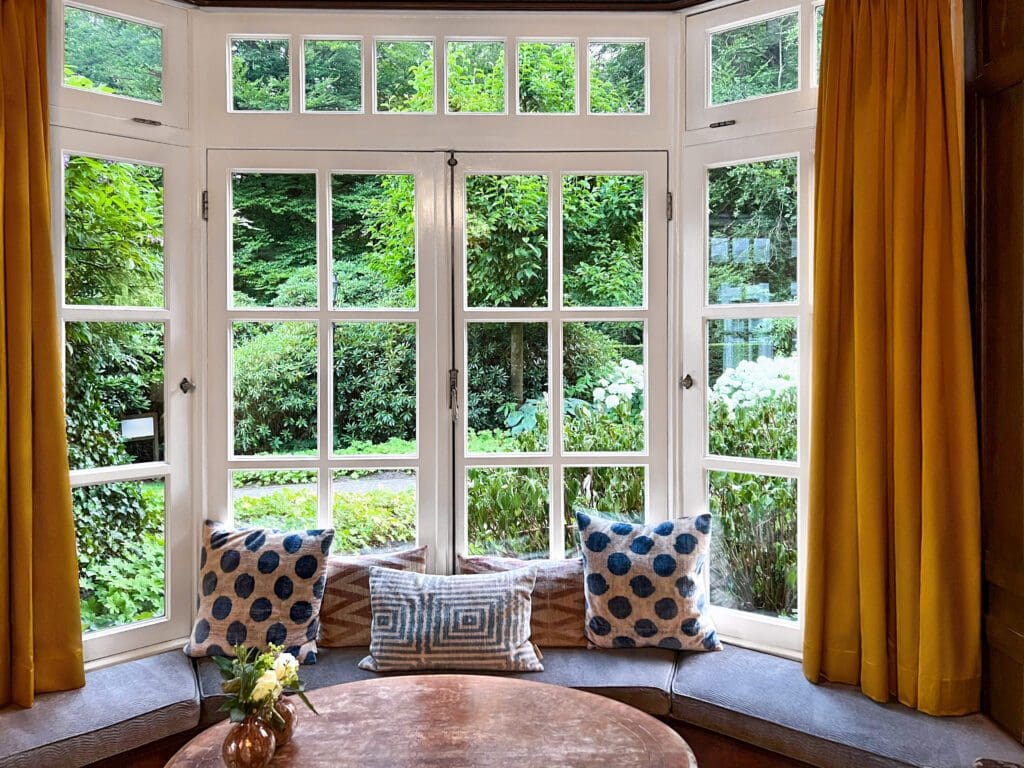 Wooden table near bay window seat with cushions in cozy room. Interior design