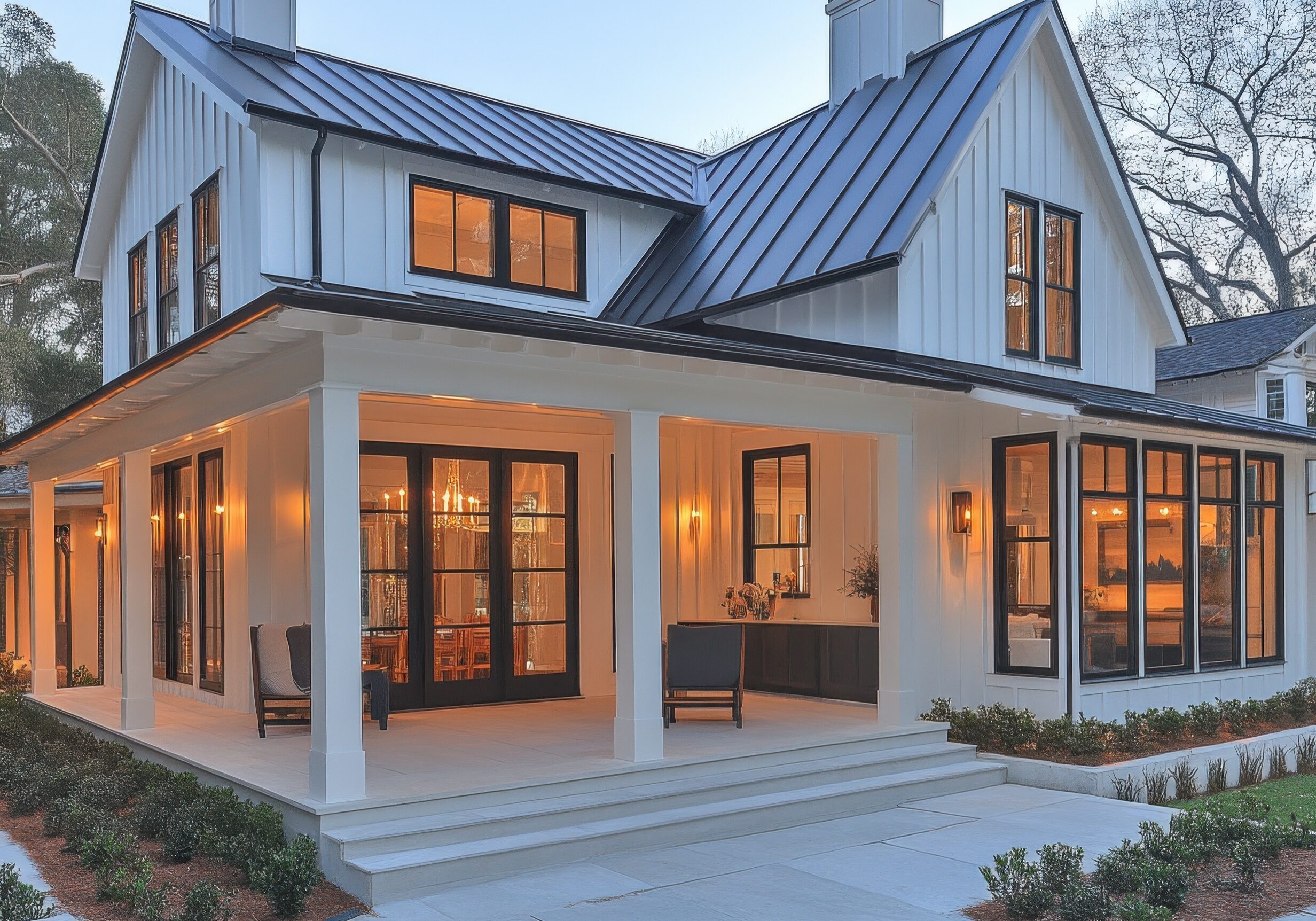 A new white contemporary farmhouse with a dark roof and large windows, featuring a cozy porch with warm lighting, surrounded by landscaping.