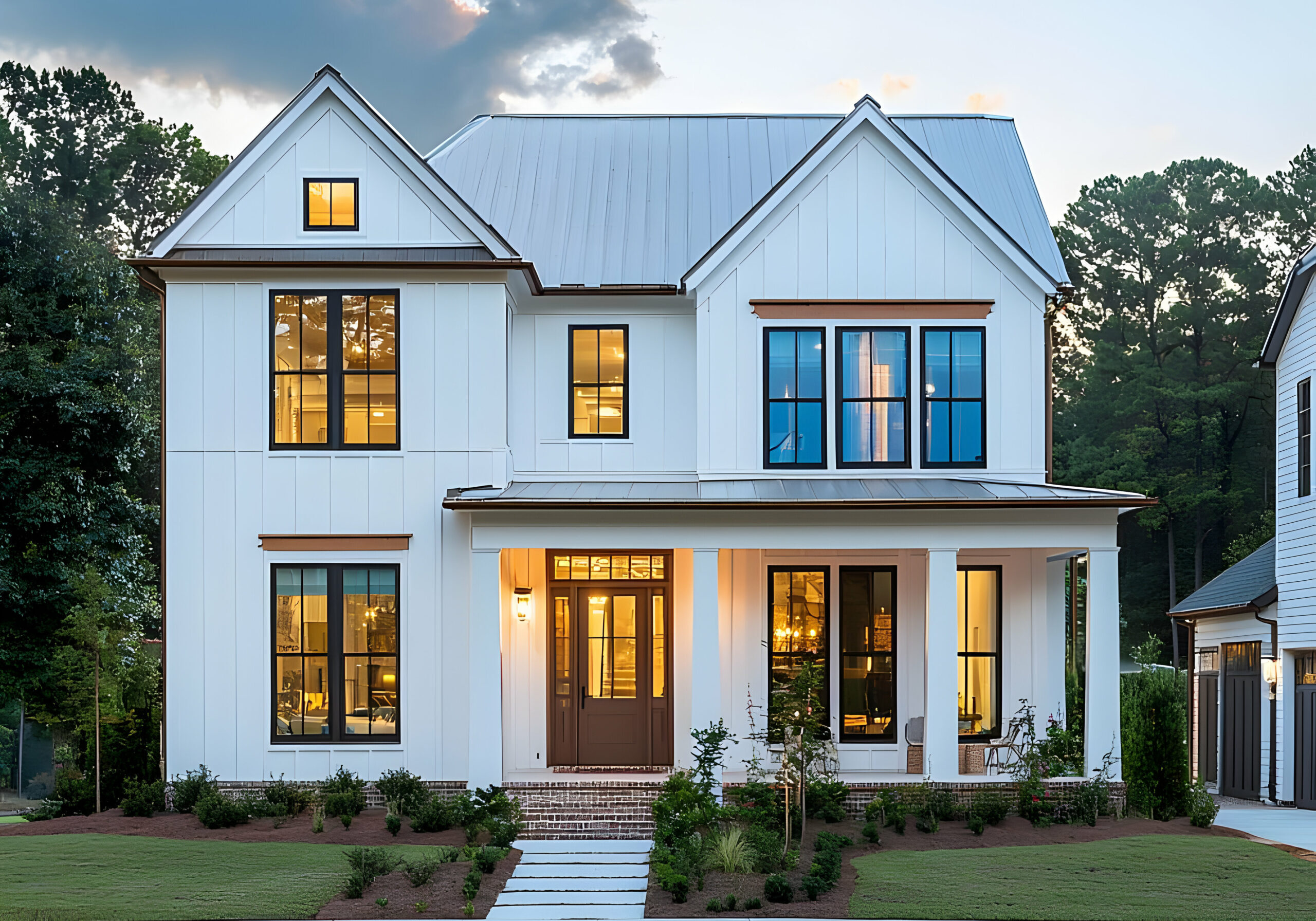 Modern white farmhouse with black-trimmed windows, metal roof, covered porch, and landscaped front yard at dusk