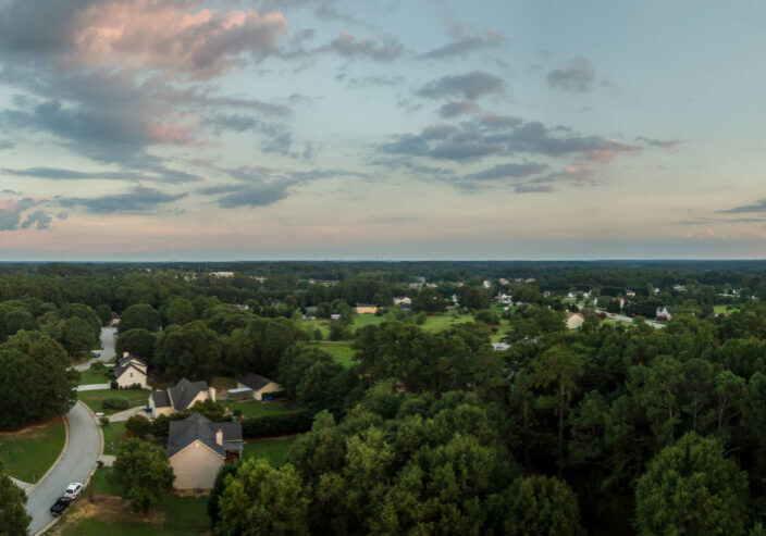 Panoramic aerial view of suburban neighborhood surrounded by dense forest at sunset with pink-tinged clouds stretching across blue sky