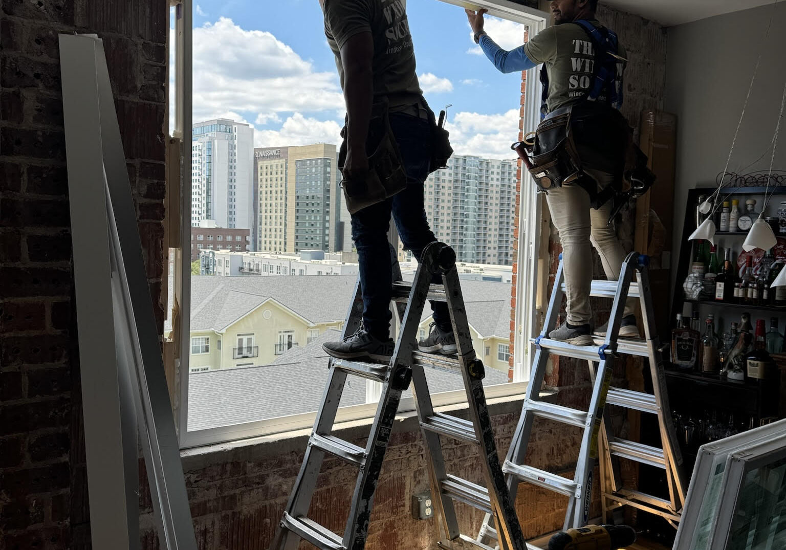 Two workers on ladders installing a large window in a brick-walled apartment with a view of downtown Atlanta, Georgia skyscrapers.